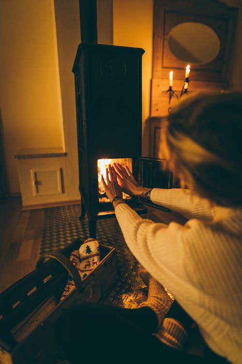 Feminine person in winter clothes warming hands close to freestanding fireplace stove